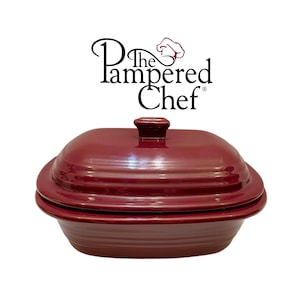 Puede incluir: Una fuente de horno rectangular de gres en color rojo oscuro profundo con una tapa a juego que tiene un asa central. El texto The Pampered Chef aparece impreso sobre la fuente en letras negras con un icono de gorro de chef rojo.