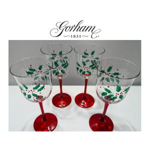 Puede incluir: Un juego de cuatro copas de vino de cristal transparente con tallos y bases rojas. Cada copa presenta un patrón pintado de hojas de acebo verdes y bayas rojas en el cáliz. El nombre de la marca Gorham 1831 aparece en texto negro sobre las copas.
