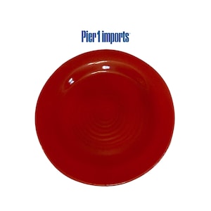 May include: A round, solid red ceramic plate with a smooth, glossy finish and a subtle concentric circular texture on the surface. The text Pier 1 imports is printed in blue above the plate, featuring a simple, clean design for dining.