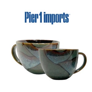 May include: Two ceramic mugs with a glossy finish featuring a dark green, blue, and brown reactive glaze pattern. The mugs are positioned in front of the blue text Pier 1 imports. The mugs have curved handles and a rounded, wide-rimmed shape.