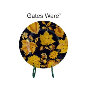 May include: A round ceramic plate featuring the text Gates Ware™ at the top. The plate has a dark navy blue background decorated with a pattern of golden yellow maple leaves and small floral elements, supported by a dark green metal display stand.