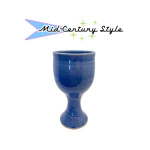 Puede incluir: Una copa de cerámica azul con un acabado liso y brillante y una base acampanada. Sobre la copa, un elemento gráfico presenta una forma de boomerang azul claro con el texto Mid-Century Style en letra cursiva negra, acentuado por pequeños símbolos de estrellas verdes.