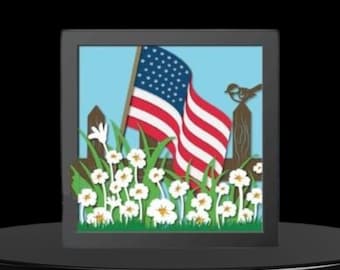 May include: A framed papercraft scene featuring an American flag waving in the wind, surrounded by white flowers and green grass. A small bird perches on a wooden fence post.