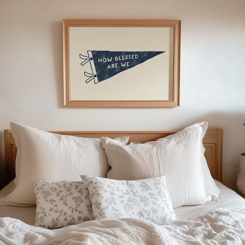 K&ouml;nnte beinhalten: Ein gerahmtes Wandkunstwerk mit dem Text "HOW BLESSED ARE WE" auf einem marineblauen Wimpel. Das Kunstwerk wird &uuml;ber einem Bett mit wei&szlig;en Kissen und floral gemusterten Wurfkissen ausgestellt. Das Bett hat ein h&ouml;lzernes Kopfteil.