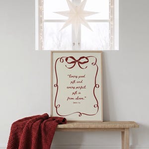 May include: A framed print with the quote "Every good gift and every perfect gift is from above." in burgundy script. A burgundy bow and decorative border frame the text. A red blanket rests on a wooden bench below the print. A white star-shaped decoration hangs in the window.