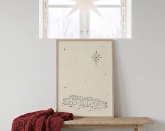 May include: A framed print with a minimalist black and white illustration of a town under a star, resting on a wooden bench. A red knitted throw blanket is draped on the bench. A star-shaped paper lantern hangs in the window.