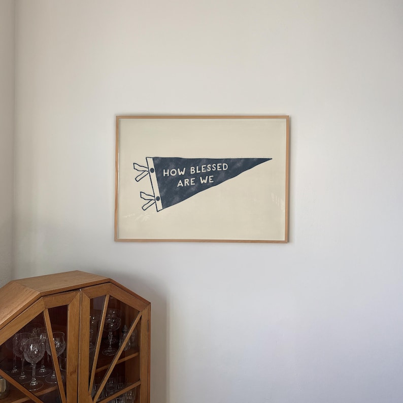 K&ouml;nnte beinhalten: Gerahmtes Wandkunstwerk in Form eines Wimpels. Der marineblaue Wimpel tr&auml;gt die wei&szlig;en Worte "HOW BLESSED ARE WE". Der Wimpel ist vor einem cremefarbenen Hintergrund platziert und in hellem Holz gerahmt.