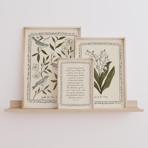 May include: A set of four framed art prints displayed on a wooden shelf. The prints feature botanical and scripture-themed designs in muted tones. One print has birds and floral elements, another has lily of the valley, and a third has text.