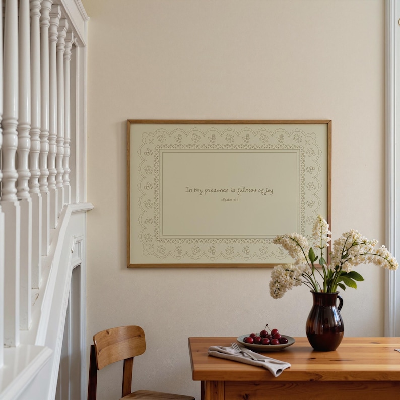May include: A framed print with the text "In thy presence is fullness of joy" hangs above a wooden table. A brown vase holds white flowers. A plate of cherries and a folded napkin sit on the table. A wooden chair is in the foreground.