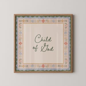 May include: A framed print featuring the text "Child of God" in a cursive font. The print has a beige background with a floral border in blue, pink, and yellow. The frame is a light brown colour.
