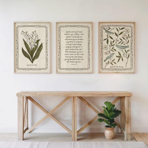 K&ouml;nnte beinhalten: Drei gerahmte Kunstdrucke mit botanischen und schriftlichen Themen. Einer zeigt eine Lilienillustration, ein anderer einen Bibelvers, und der dritte zeigt V&ouml;gel zwischen Zweigen. Die Rahmen sind hell und die Drucke werden &uuml;ber einem Holztisch ausgestellt.