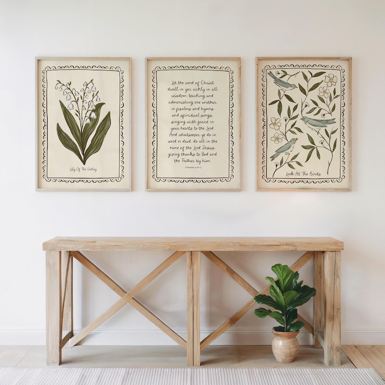 K&ouml;nnte beinhalten: Drei gerahmte Kunstdrucke mit botanischen und schriftlichen Themen. Einer zeigt eine Lilienillustration, ein anderer einen Bibelvers, und der dritte zeigt V&ouml;gel zwischen Zweigen. Die Rahmen sind hell und die Drucke werden &uuml;ber einem Holztisch ausgestellt.