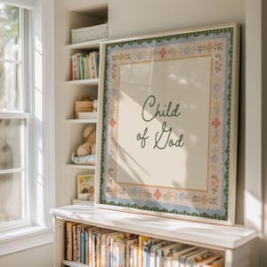 Könnte beinhalten: Gerahmter Kunstdruck mit dem Schriftzug "Child of God" in grüner Schrift. Der Druck hat einen cremefarbenen Hintergrund und einen dekorativen Blumenrand in Blau, Rosa und Gelb. Der Rahmen ist weiß und steht auf einem Regal.