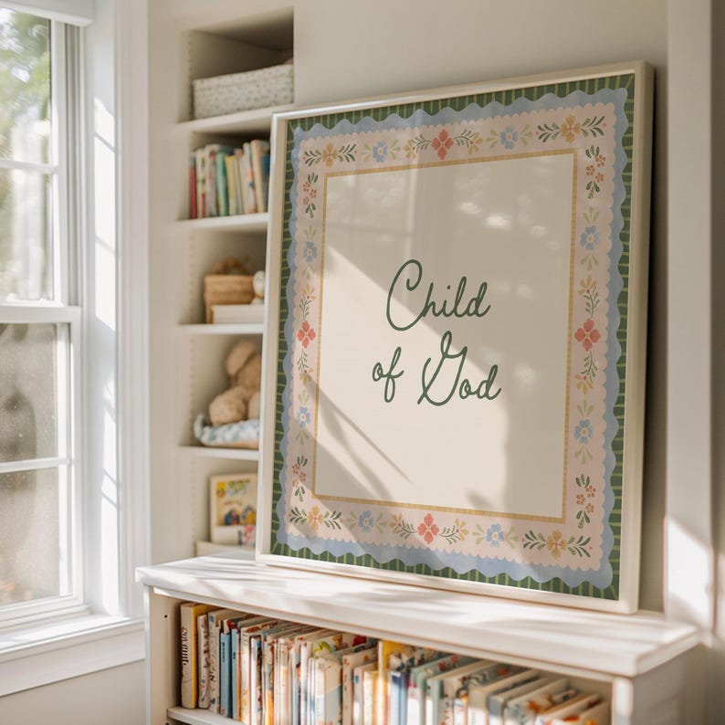 May include: A framed art print featuring the text "Child of God" in green script. The print has a cream background and a decorative floral border in blue, pink, and yellow. The frame is white, and the print is displayed on a shelf.