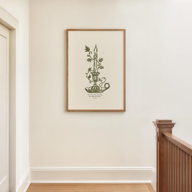 May include: A framed print with a green illustration of a candle in a decorative holder with leafy vines. The artwork includes text at the bottom. The frame is light brown, and the print hangs on a cream-coloured wall.