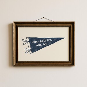 K&ouml;nnte beinhalten: Gerahmter Druck mit einem marineblauen Wimpel mit dem wei&szlig;en Text "HOW BLESSED ARE WE". Der Wimpel befindet sich vor einem cremefarbenen Hintergrund und ist in einem dunkelbraunen Holzrahmen eingefasst.