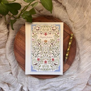 May include: A Prayer Journal with a floral design, birds, and the words "Prayer Journal" in script. The journal is on a wooden tray with a gold pen. The journal has a light blue border.