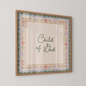 May include: A framed print with the words "Child of God" in cursive script. The artwork has a light beige background and a floral border in pink, blue, and green. The frame is made of wood.