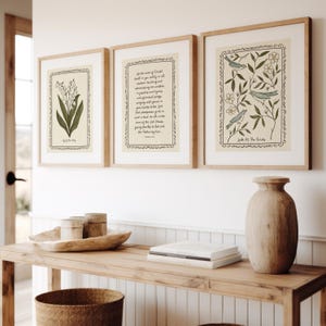 K&ouml;nnte beinhalten: Drei gerahmte Kunstdrucke mit botanischen und inspirierenden Themen. Die Drucke zeigen eine Maigl&ouml;ckchen-Illustration, einen Bibelvers und ein Vogeldesign. Die Rahmen sind hellbraun und die Drucke haben einen cremefarbenen Hintergrund.