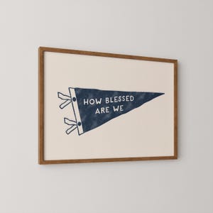 K&ouml;nnte beinhalten: Gerahmter Druck mit einem marineblauen Wimpel mit dem wei&szlig;en Text "HOW BLESSED ARE WE". Der Wimpel befindet sich vor einem cremefarbenen Hintergrund und ist in einem warmbraunen Holzrahmen eingefasst.