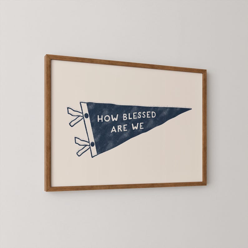 K&ouml;nnte beinhalten: Gerahmter Druck mit einem marineblauen Wimpel mit dem wei&szlig;en Text "HOW BLESSED ARE WE". Der Wimpel befindet sich vor einem cremefarbenen Hintergrund und ist in einem warmbraunen Holzrahmen eingefasst.