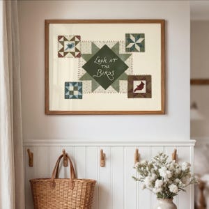 May include: A framed artwork featuring a central green starburst design with the text "Look at the Birds." The artwork includes four smaller, square quilt-like patches with bird and geometric patterns. The frame is a warm brown color.