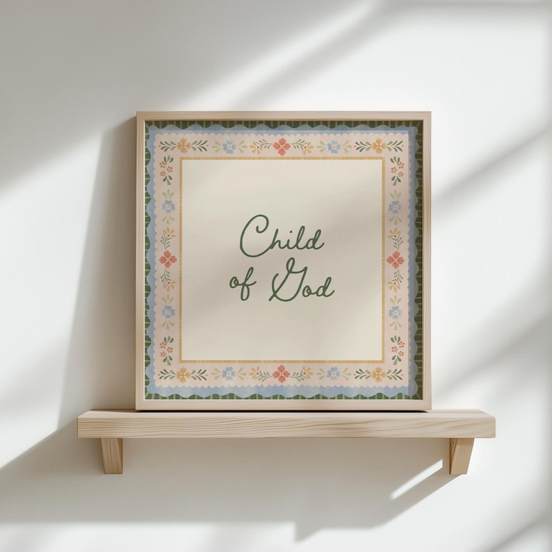 May include: Square framed art print with the text "Child of God" in a script font. The print has a cream-coloured background with a floral border in pastel colours. The frame is a light wood colour.