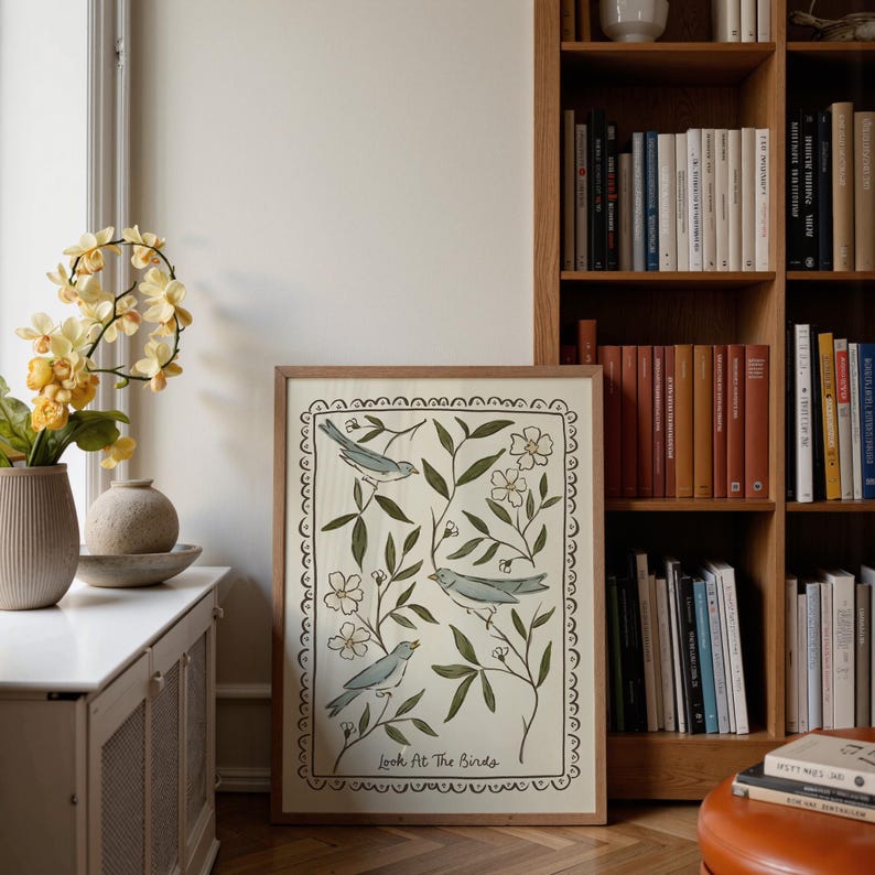 Pu&ograve; includere: Una stampa artistica incorniciata con tre uccelli blu tra foglie verdi e fiori bianchi. L'opera d'arte &egrave; bordata da un bordo decorativo e include il testo "Look At The Birds" in basso. La stampa &egrave; esposta vicino a una finestra e a una libreria.