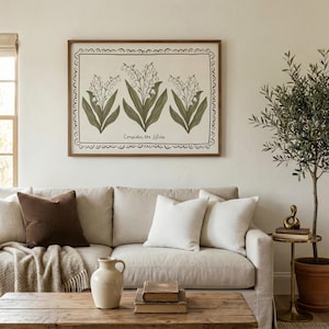 Peut inclure: Un cadre avec une illustration botanique de trois brins de muguet verts et blancs, avec le texte "Consider the lilies". L'œuvre est au-dessus d'un canapé beige avec des coussins. Une table basse en bois et un petit arbre en pot sont au premier plan.