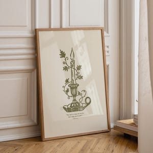 May include: A framed print with a green illustration of a candle in a decorative holder, adorned with leafy vines. The artwork is set against a cream background and includes a quote at the bottom. The frame is light brown.