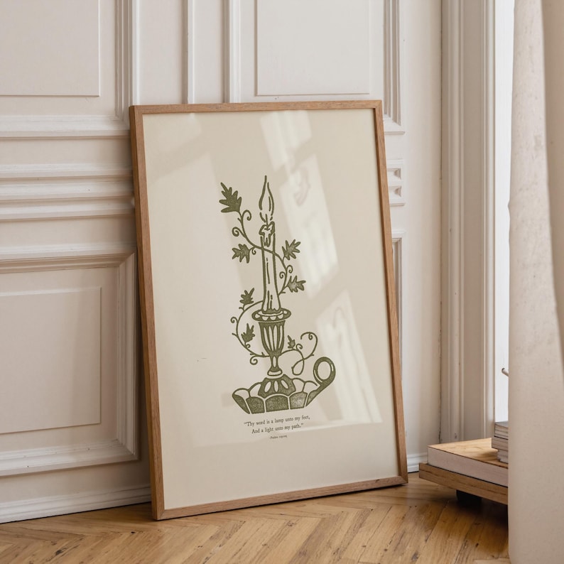 May include: A framed print with a green illustration of a candle in a decorative holder, adorned with leafy vines. The artwork is set against a cream background and includes a quote at the bottom. The frame is light brown.