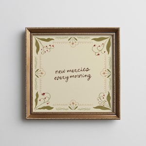 May include: Square framed art print with the text "new mercies every morning" in a handwritten style. The print has a cream background with floral corner designs in red, green, and cream. The frame is a dark gold color.