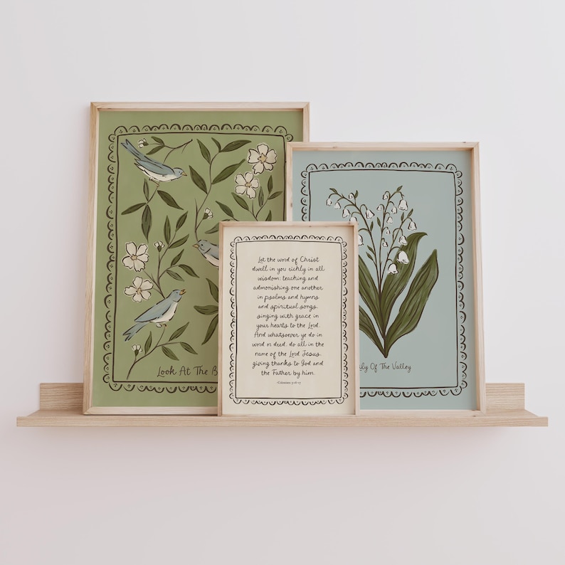 May include: Four framed art prints displayed on a wooden shelf. The prints feature botanical and scripture-themed designs. One print has birds and floral elements, another has lily of the valley, and a third has text. The frames are light wood.