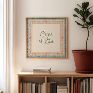 May include: A framed print featuring the text "Child of God" in a cursive font. The print has a cream-colored background and a decorative border with floral and scalloped details in blue, green, and peach. The frame is a light wood colour.
