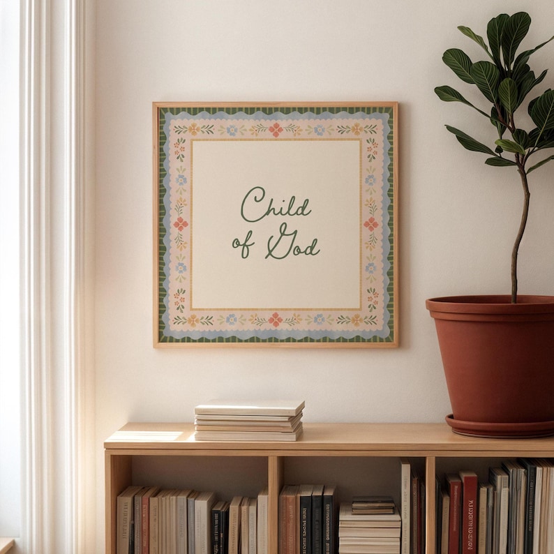 May include: A framed print featuring the text "Child of God" in a cursive font. The print has a cream-colored background and a decorative border with floral and scalloped details in blue, green, and peach. The frame is a light wood colour.