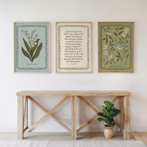 May include: Three framed art prints with botanical and scripture themes. The prints feature a lily of the valley, a Bible verse, and birds with floral elements. The frames are light-coloured, and the prints are displayed above a wooden console table.