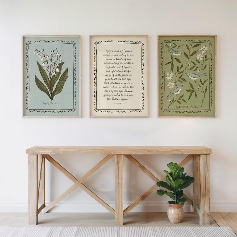 May include: Three framed art prints with botanical and scripture themes. The prints feature a lily of the valley, a Bible verse, and birds with floral elements. The frames are light-coloured, and the prints are displayed above a wooden console table.