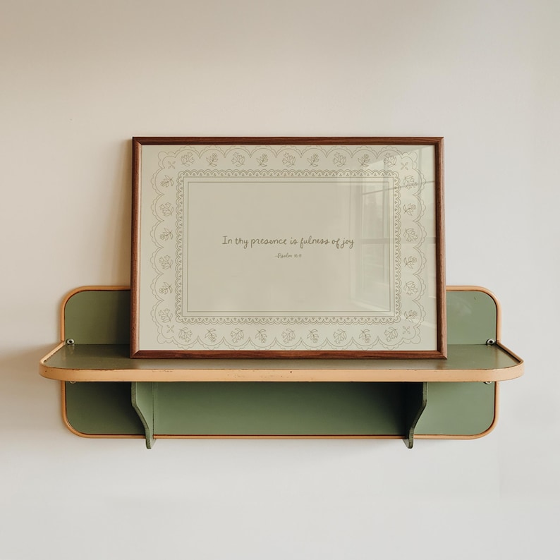 May include: A framed print with the text "In thy presence is fullness of joy" displayed on a green and tan shelf. The frame is brown and the print has a decorative border.