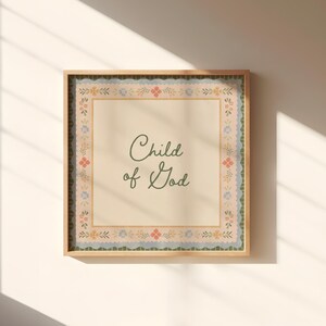 May include: A framed print featuring the words "Child of God" in a cursive font. The print has a beige background with a floral border in pink, blue, and green. The frame is a light brown colour.