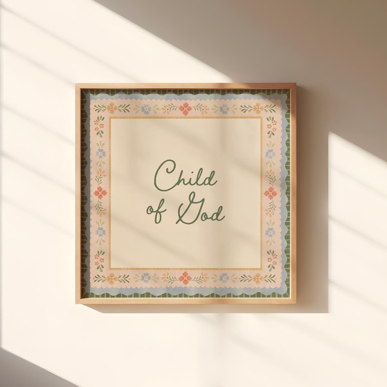 May include: A framed print featuring the words "Child of God" in a cursive font. The print has a beige background with a floral border in pink, blue, and green. The frame is a light brown colour.