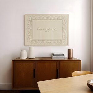 May include: A framed print with the text "In thy presence is fullness of joy" in a decorative frame. The print hangs above a wooden cabinet with two white vases, a brown vase, and a stack of books. The cabinet is in a room with a wooden table.