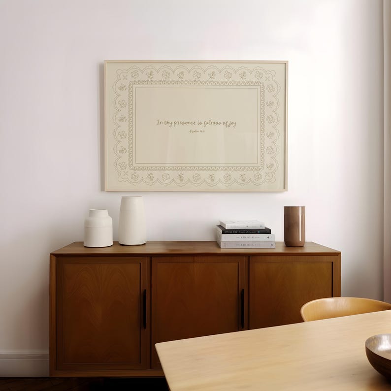 May include: A framed print with the text "In thy presence is fullness of joy" in a decorative frame. The print hangs above a wooden cabinet with two white vases, a brown vase, and a stack of books. The cabinet is in a room with a wooden table.