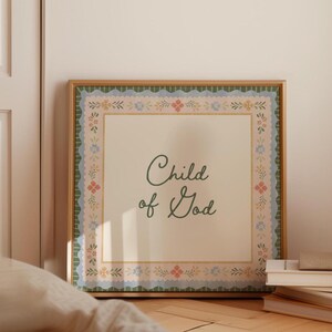 May include: A framed print with the words "Child of God" in elegant script. The artwork features a cream-coloured background with a decorative border of floral and scalloped patterns in shades of green, pink, and blue. The frame is a warm, natural wood tone.