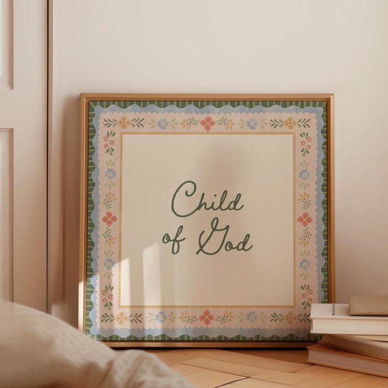 May include: A framed print with the words "Child of God" in elegant script. The artwork features a cream-coloured background with a decorative border of floral and scalloped patterns in shades of green, pink, and blue. The frame is a warm, natural wood tone.