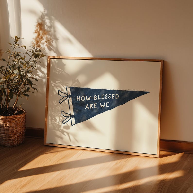 K&ouml;nnte beinhalten: Gerahmter Druck mit einem marineblauen Wimpel mit dem wei&szlig;en Text "HOW BLESSED ARE WE". Der Wimpel hat eine dreieckige Form mit wei&szlig;en Akzenten. Der Rahmen ist hellbraun.