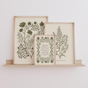 May include: Three framed botanical art prints with green floral illustrations and a light beige background. The middle print has text that reads "The grass withereth, the flower fadeth, but the word of our God shall stand forever."