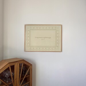 May include: A framed print with the text "In thy presence is fullness of joy" in a decorative frame. The print has a beige background with a detailed border. The frame is a light brown color.