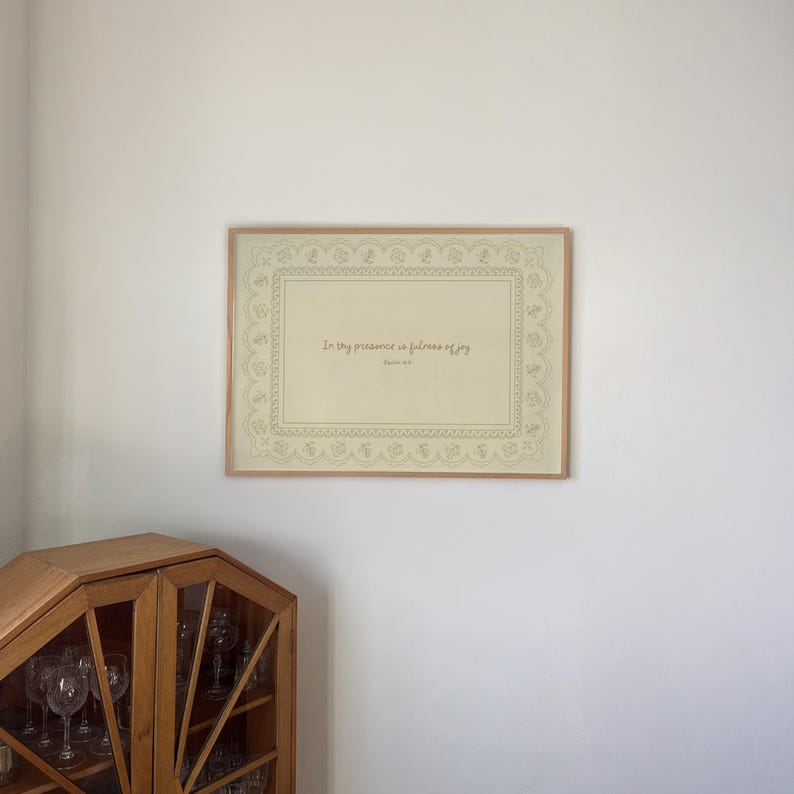 May include: A framed print with the text "In thy presence is fullness of joy" in a decorative frame. The print has a beige background with a detailed border. The frame is a light brown color.