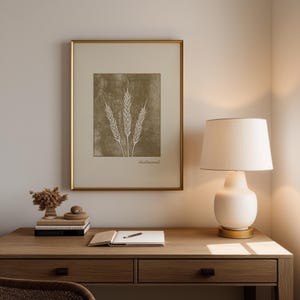 Op de afbeelding: Een ingelijste print met een tarwe-illustratie in een gouden lijst. Het kunstwerk heeft een neutraal kleurenpalet met een beige achtergrond en tarwearen in een lichtere tint. Het woord "Redeemed" staat onderaan.