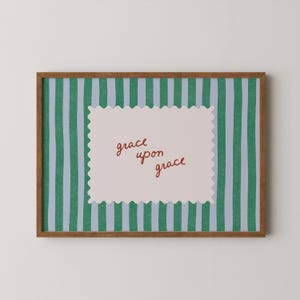 May include: A framed print with a brown wooden frame. The background features vertical stripes in green and light blue. A light pink, scalloped-edge rectangle in the center displays the words "grace upon grace" in a handwritten style.